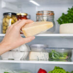 Hand holding a wedge of parmesan cheese inside a clean, well-lit refrigerator, with neatly arranged jars, eggs, greens, and vegetables on white shelves in the background.