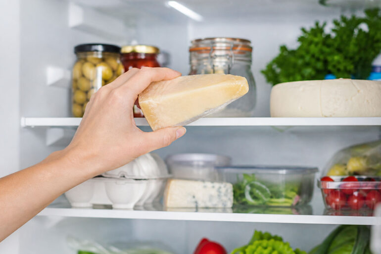 Hand holding a wedge of parmesan cheese inside a clean, well-lit refrigerator, with neatly arranged jars, eggs, greens, and vegetables on white shelves in the background.