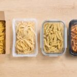 Overhead flat lay on a light wood surface. Four distinct items arranged left to right: an open box of dry penne, a sealed refrigerated fresh pasta package, a glass container of cooked pasta with a tight lid, and a pre-made refrigerated pasta meal tray with a use-by date visible in 2026. Each item clearly separated