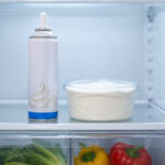 an open refrigerator interior. A can of aerosol whipped cream stands upright on the middle interior shelf, centered in frame. A sealed tub of whipped topping sits beside it on the same shelf