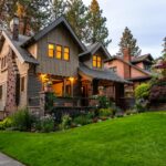 A beautifully lit Victorian-style house surrounded by a lush garden at twilight, showcasing classic architecture.