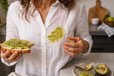 A woman in a white linen shirt stands in a kitchen holding avocado toast after accidentally smearing avocado across the front of her shirt, with a cut avocado, lemon wedges, and seasoning visible on the counter nearby.
