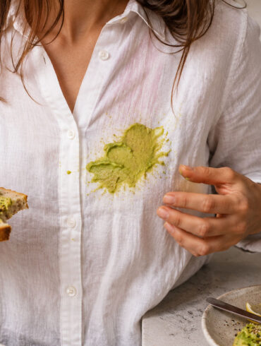 A woman in a white linen shirt stands in a kitchen holding avocado toast after accidentally smearing avocado across the front of her shirt, with a cut avocado, lemon wedges, and seasoning visible on the counter nearby.