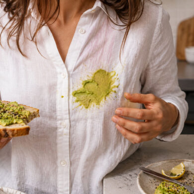 A woman in a white linen shirt stands in a kitchen holding avocado toast after accidentally smearing avocado across the front of her shirt, with a cut avocado, lemon wedges, and seasoning visible on the counter nearby.