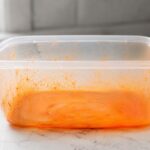 A close-up of a clear plastic food storage container with a vivid orange-stained interior, sitting on a white kitchen counter.