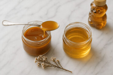 Two open jars of honey side by side. The left jar is manuka. The right jar is lighter, more golden and clearly more fluid — regular clover honey. A single silver spoon rests across the manuka jar with a slow dark drip. A small bear-shaped honey bottle sits slightly behind and out of focus to the right, suggesting the supermarket alternative. One dried manuka flower sprig placed loosely between the jars