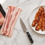 Overhead flat lay on a white marble surface. Left side: an open package of raw bacon with three or four strips pulled out and fanned slightly, showing the pink meat and white fat. Right side: a small white plate with four or five strips of cooked crispy bacon. Center: a knife resting at an angle on the board