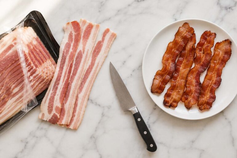 Overhead flat lay on a white marble surface. Left side: an open package of raw bacon with three or four strips pulled out and fanned slightly, showing the pink meat and white fat. Right side: a small white plate with four or five strips of cooked crispy bacon. Center: a knife resting at an angle on the board