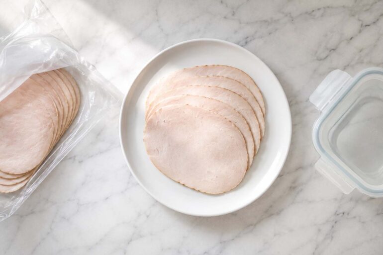 A sealed package of deli turkey sits upright on the middle interior shelf, centered in frame. A small airtight glass container of sliced turkey sits beside it.