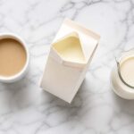 Flat lay on white marble. Center: open half and half carton, slightly tilted. Left: white coffee mug. Right: small glass pitcher of cream.