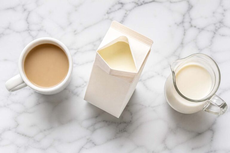 Flat lay on white marble. Center: open half and half carton, slightly tilted. Left: white coffee mug. Right: small glass pitcher of cream.