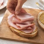Overhead or slight angle kitchen counter scene. A simple sandwich being assembled with sliced ham as the focus. One slice of bread open-faced with several folded slices of pink ham being placed on top by a hand. The second slice of bread sits nearby