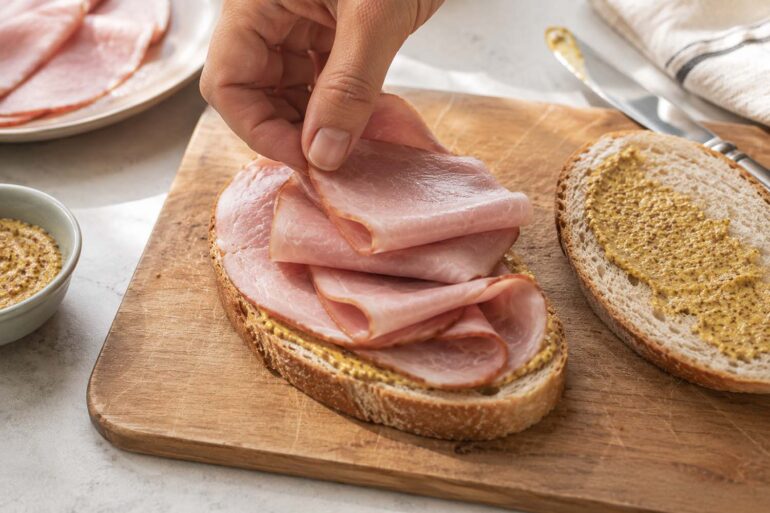 Overhead or slight angle kitchen counter scene. A simple sandwich being assembled with sliced ham as the focus. One slice of bread open-faced with several folded slices of pink ham being placed on top by a hand. The second slice of bread sits nearby
