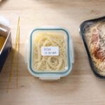 Overhead flat lay on a light wood surface. Left side: an open box of dry spaghetti with a few loose strands beside it. Center: a glass container of leftover cooked pasta with a tight-fitting lid and a handwritten date label. Right side: a store-bought refrigerated pasta meal package