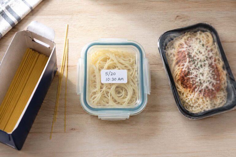 Overhead flat lay on a light wood surface. Left side: an open box of dry spaghetti with a few loose strands beside it. Center: a glass container of leftover cooked pasta with a tight-fitting lid and a handwritten date label. Right side: a store-bought refrigerated pasta meal package