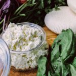 a container of ricotta cheese on a wooden board with greens and wheels of ricotta