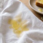 Photorealistic close-up of a fresh melted butter stain spreading across a white linen shirt, the fabric visibly absorbing the butter