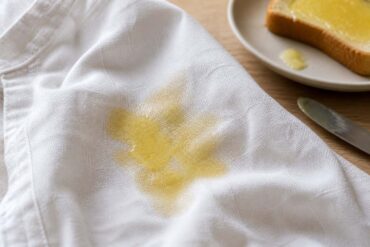 Photorealistic close-up of a fresh melted butter stain spreading across a white linen shirt, the fabric visibly absorbing the butter