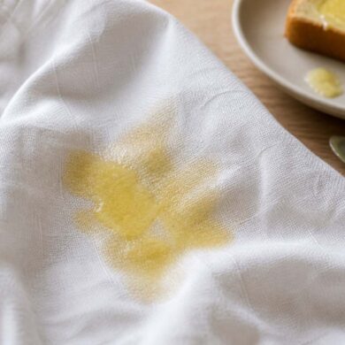 Photorealistic close-up of a fresh melted butter stain spreading across a white linen shirt, the fabric visibly absorbing the butter