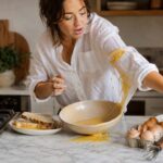 A woman in her 30s wearing a white linen shirt stands at a marble kitchen counter making French toast as beaten egg mixture spills from a shallow bowl onto her sleeve. Slices of bread, cracked eggs, a whisk, and a skillet sit nearby in a warm naturally lit home kitchen scene.