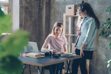 Two women working together at a desk in a modern, plant-filled office.