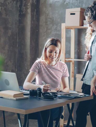Two women working together at a desk in a modern, plant-filled office.