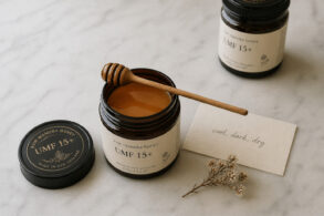 Overhead flat lay on honed white marble. A single open jar of dark amber manuka honey, lid resting beside it, a dry wooden honey spoon across the top. Beside it a small handwritten card reading "cool, dark, dry" in minimal script, slightly angled. One dried manuka flower sprig placed loosely nearby. A second sealed jar slightly out of focus in the background suggesting long-term storage.