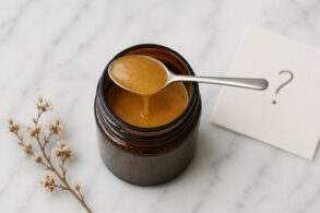 Overhead view of an open amber glass jar filled with thick, golden-brown manuka honey on a white marble surface. A small stainless steel spoon rests across the jar, with a slow drip of honey falling back inside. To the right, a slightly out-of-focus off-white card shows a handwritten question mark. To the left, a delicate dried flower sprig lies on the marble.