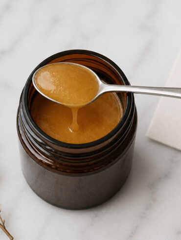 Overhead view of an open amber glass jar filled with thick, golden-brown manuka honey on a white marble surface. A small stainless steel spoon rests across the jar, with a slow drip of honey falling back inside. To the right, a slightly out-of-focus off-white card shows a handwritten question mark. To the left, a delicate dried flower sprig lies on the marble.