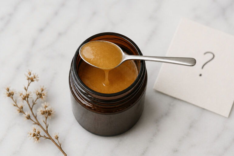 Overhead view of an open amber glass jar filled with thick, golden-brown manuka honey on a white marble surface. A small stainless steel spoon rests across the jar, with a slow drip of honey falling back inside. To the right, a slightly out-of-focus off-white card shows a handwritten question mark. To the left, a delicate dried flower sprig lies on the marble.