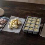 Dark wood surface. Four distinct stations arranged naturally: far left, a whole head of garlic beside several unpeeled cloves going into a zip-top freezer bag. Center left, a small pile of peeled cloves on a sheet of parchment. Center right, a silicone ice cube tray with minced garlic portioned into each cavity, a small spoon resting beside it. Far right, two frozen garlic-oil cubes on the parchment with a small label reading "keep frozen."