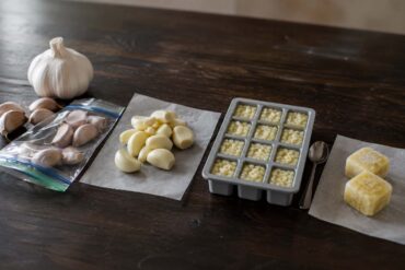 Dark wood surface. Four distinct stations arranged naturally: far left, a whole head of garlic beside several unpeeled cloves going into a zip-top freezer bag. Center left, a small pile of peeled cloves on a sheet of parchment. Center right, a silicone ice cube tray with minced garlic portioned into each cavity, a small spoon resting beside it. Far right, two frozen garlic-oil cubes on the parchment with a small label reading "keep frozen."