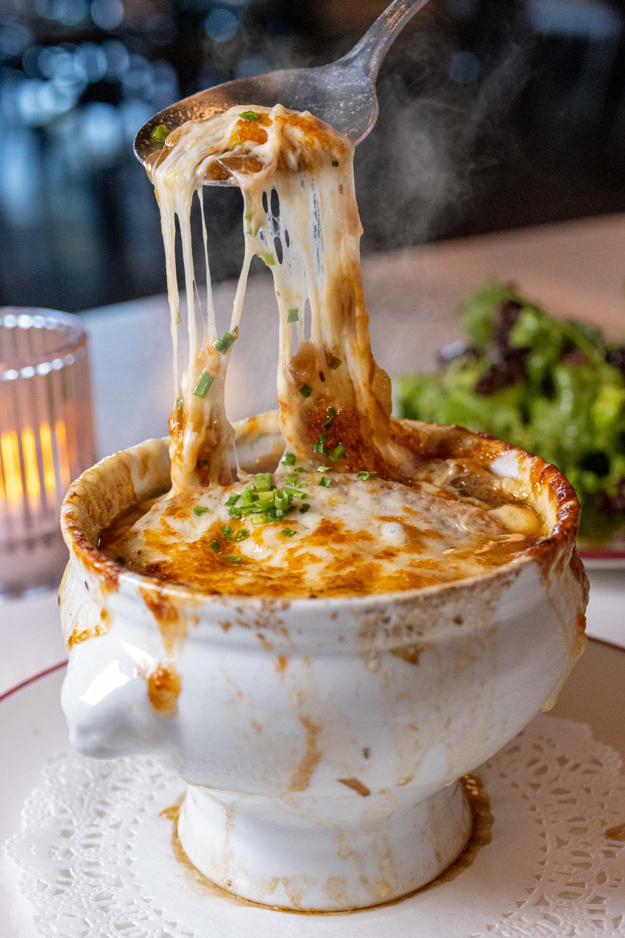 Classic French onion soup in a white crock with a dramatic cheese pull, garnished with fresh chives, served with a mixed green salad at Chez Frites