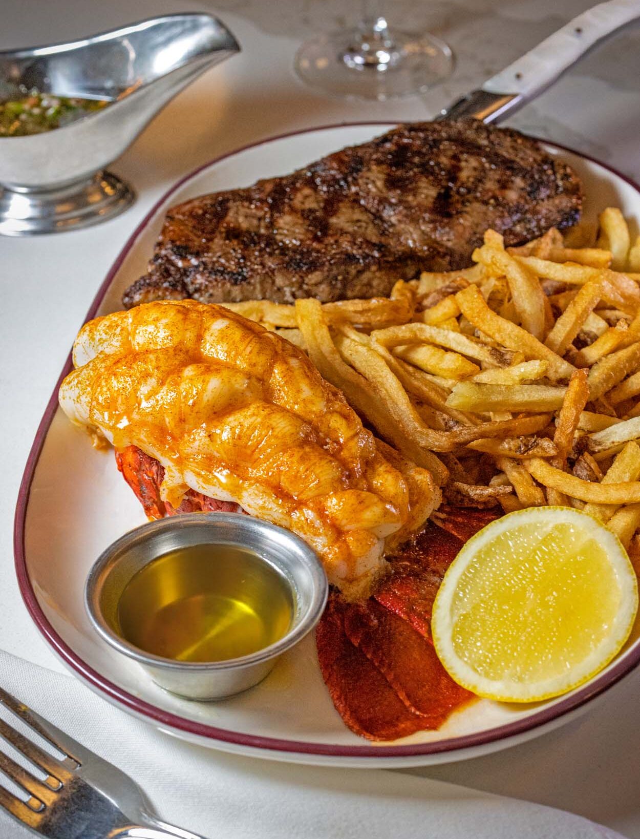 Surf and Turf Frites special at Chez Frites featuring a prime NY strip steak and South African lobster tail with golden hand-cut fries, drawn butter, and lemon on a white plate with burgundy rim