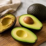 Fresh ripe avocados on a rustic wooden board, including one halved avocado with the pit and another halved avocado showing creamy green flesh, styled on a neutral linen backdrop