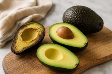 Fresh ripe avocados on a rustic wooden board, including one halved avocado with the pit and another halved avocado showing creamy green flesh, styled on a neutral linen backdrop