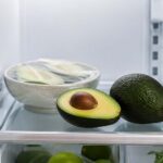 A clean, modern refrigerator interior with the door open, viewed at eye level. On the middle shelf, a halved ripe avocado with vibrant green flesh sits beside a whole dark-green Hass avocado. In the background, a small bowl containing avocado halves wrapped in clear reusable food wrap suggests refrigerated storage after cutting.