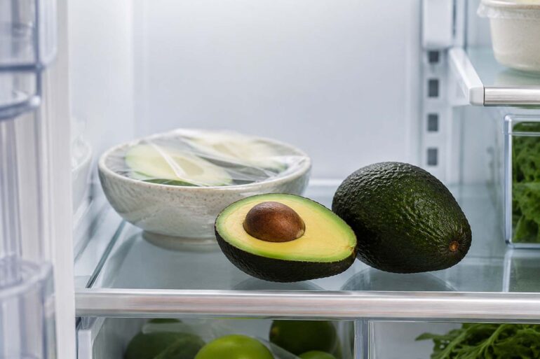 A clean, modern refrigerator interior with the door open, viewed at eye level. On the middle shelf, a halved ripe avocado with vibrant green flesh sits beside a whole dark-green Hass avocado. In the background, a small bowl containing avocado halves wrapped in clear reusable food wrap suggests refrigerated storage after cutting.