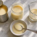 a can of sweetened condensed milk with its top opened beside a small glass jar of the thick, pale golden milk poured out, a spoon resting in a small bowl showing the syrupy, dense texture, and a measuring cup nearby.