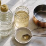 o corn syrup bottle with its cap open beside a small glass jar of the thick, pale golden syrup mid-pour, a spoon resting in a small dish showing the syrup's smooth viscous texture, and a small saucepan nearby