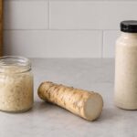 A jar of prepared horseradish, a fresh horseradish root, and a bottle of creamy horseradish sauce on a kitchen counter