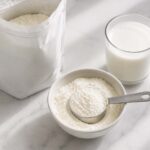 Does Powdered Milk Go Bad? A plain unlabeled box or bag of fine white milk powder beside it, a small white ceramic bowl filled with the powder and a dry measuring cup resting in it, and a glass of reconstituted white milk sitting nearby.