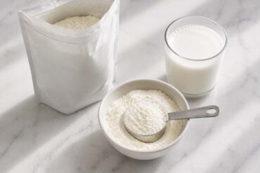 Does Powdered Milk Go Bad? A plain unlabeled box or bag of fine white milk powder beside it, a small white ceramic bowl filled with the powder and a dry measuring cup resting in it, and a glass of reconstituted white milk sitting nearby.