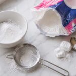 a partially open bag of powdered sugar beside a small white ceramic bowl dusted with fine white powder, a fine mesh sifter with a light dusting of powdered sugar sifted beneath it, and two or three small soft lumps of sugar beside a spoon used to break them up