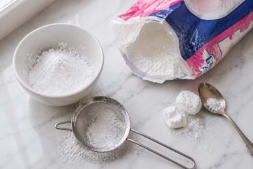 a partially open bag of powdered sugar beside a small white ceramic bowl dusted with fine white powder, a fine mesh sifter with a light dusting of powdered sugar sifted beneath it, and two or three small soft lumps of sugar beside a spoon used to break them up