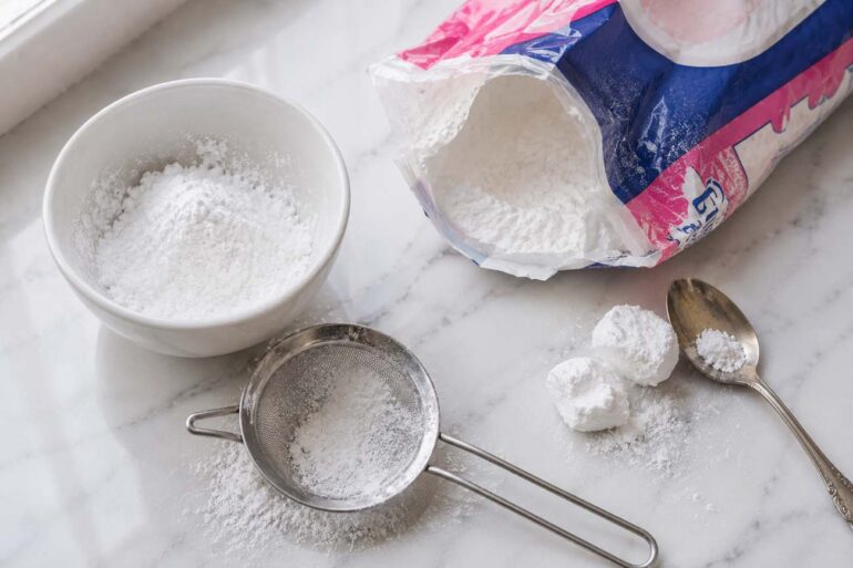 a partially open bag of powdered sugar beside a small white ceramic bowl dusted with fine white powder, a fine mesh sifter with a light dusting of powdered sugar sifted beneath it, and two or three small soft lumps of sugar beside a spoon used to break them up