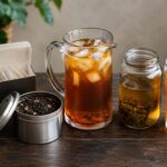 Five forms of tea on a dark wood surface: sealed tea bags in a box, loose leaf tea in an open tin, a glass pitcher of brewed iced tea, a jar of cold brew tea, and a glass of sun tea with lemon