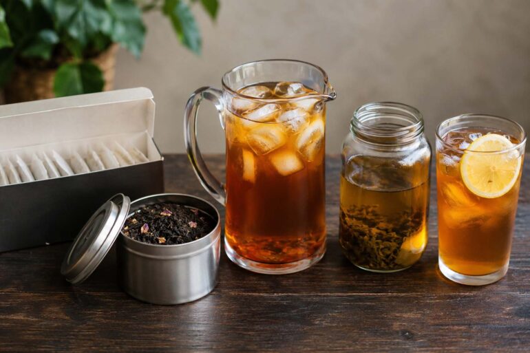 Five forms of tea on a dark wood surface: sealed tea bags in a box, loose leaf tea in an open tin, a glass pitcher of brewed iced tea, a jar of cold brew tea, and a glass of sun tea with lemon