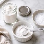 a small glass spice jar of white cream of tartar powder with its lid beside it, a white ceramic ramekin with a small mound of the powder and a measuring spoon resting in it, and a second small bowl beside it showing a vigorous white fizz from the baking soda water test