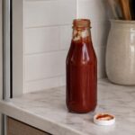 An opened bottle of ketchup on a kitchen counter next to a refrigerator.
